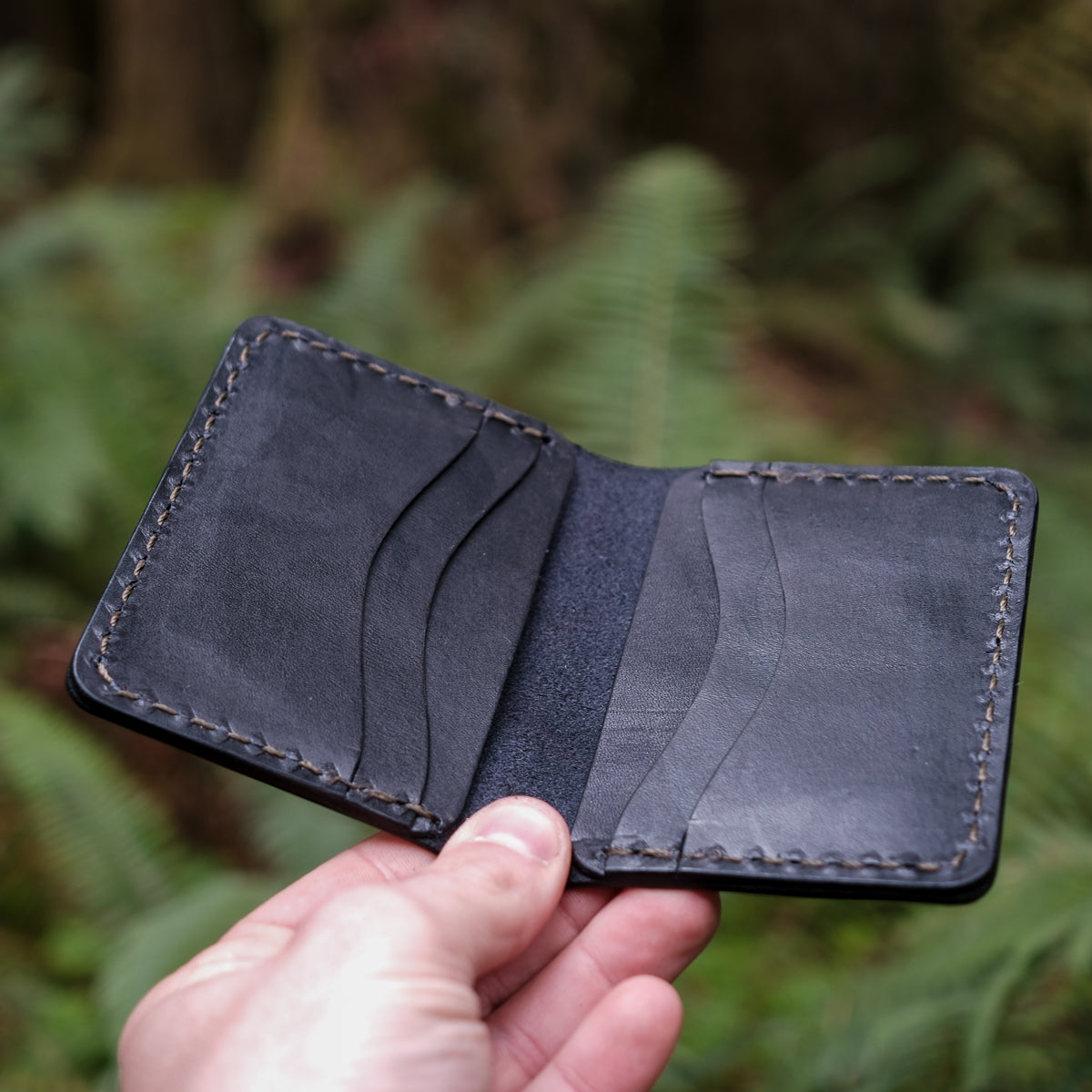 Hand holding a black leather wallet against a blurred green foliage background